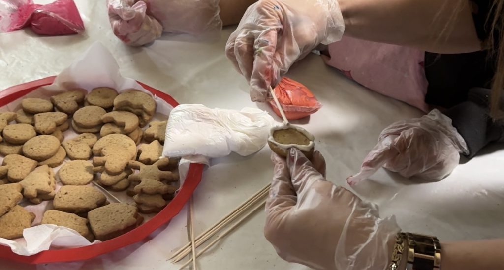 Ruka u zaštitnoj rukavici precizno nanosi belu glazuru na pečeni medenjak pomoću drvenog štapića.