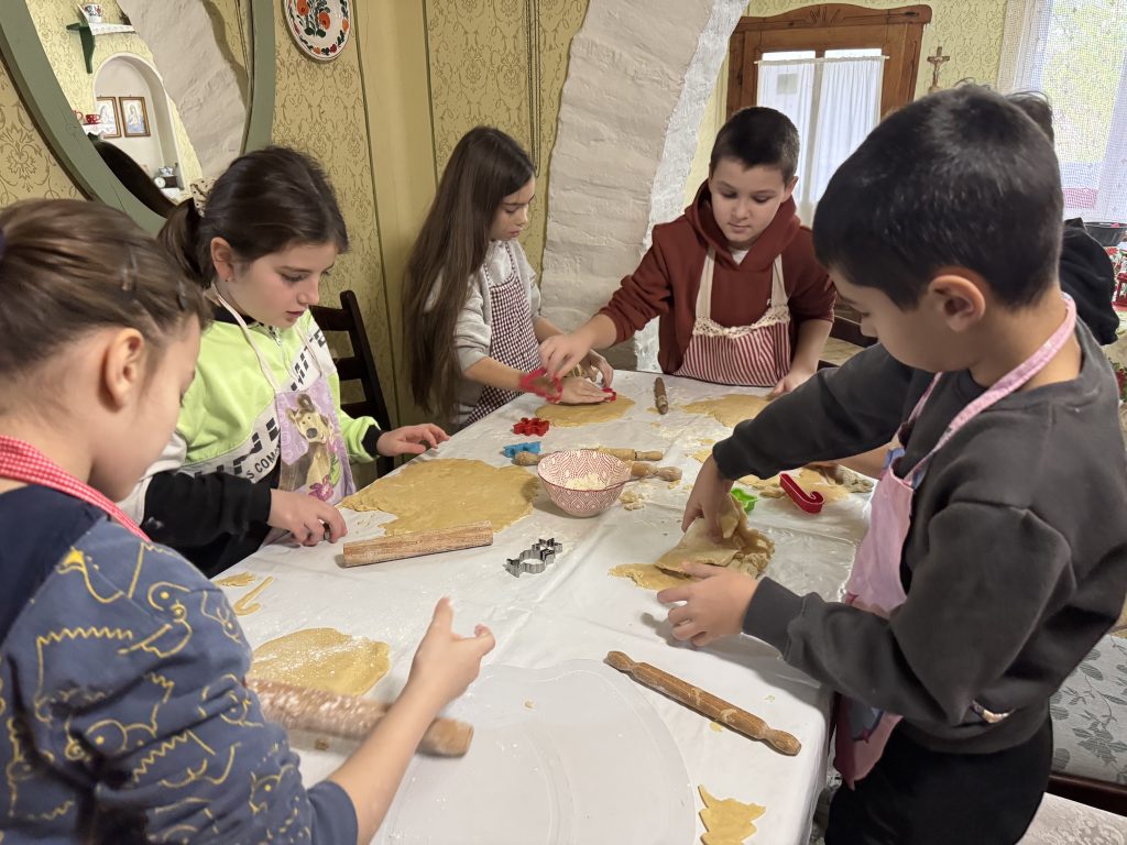 Deca za stolom mese testo za domaće medenjake koristeći drvene oklagije i brašno.