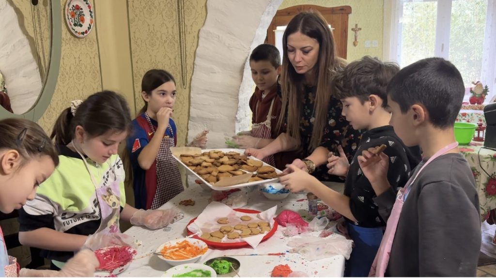 Žena pomaže grupi dece oko stola dok zajedno pripremaju i ređaju medenjake.