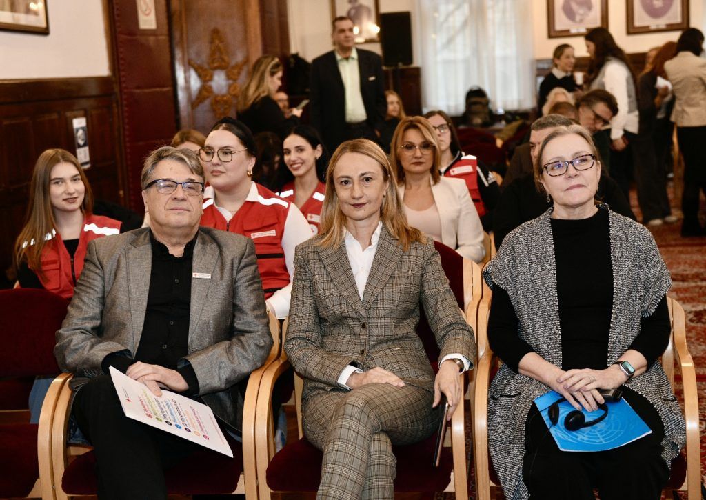 Tri osobe u prvom planu sede u publici, okružene drugim učesnicima. S leva na desno: muškarac u sivom sakou, žena u kariranom odelu i žena u crno-beloj pletenoj tunici. U pozadini su vidljivi volonteri Crvenog krsta u crvenim prslucima.