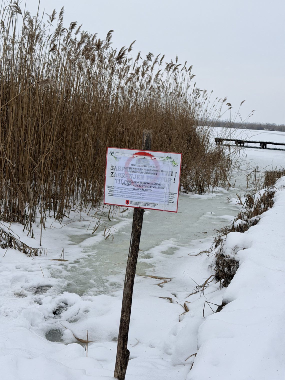 Tabla sa upozorenjem na srpskom i mađarskom jeziku stoji pored trske i obale, dok su u pozadini zaleđena jezera i drveni mol.