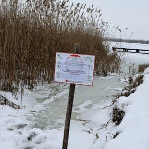 Tabla sa upozorenjem na srpskom i mađarskom jeziku stoji pored trske i obale, dok su u pozadini zaleđena jezera i drveni mol.
